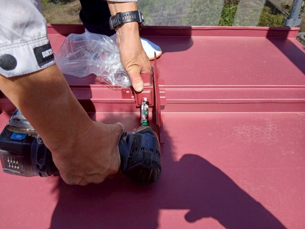 金属屋根の雪止め金具を固定している作業の様子。電動ドライバーを使用して新しい金具を取り付けている。